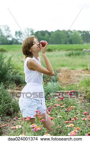 Picture - Young woman standing in flower garden, smelling apple, eyes closed. Fotosearch