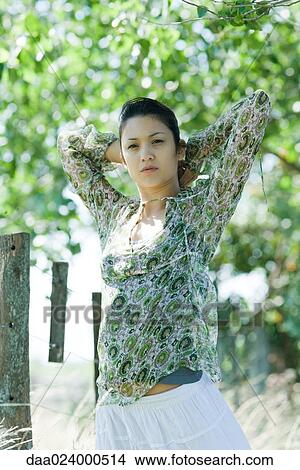 Picture - Young woman standing in rural setting, hands behind head. Fotosearch