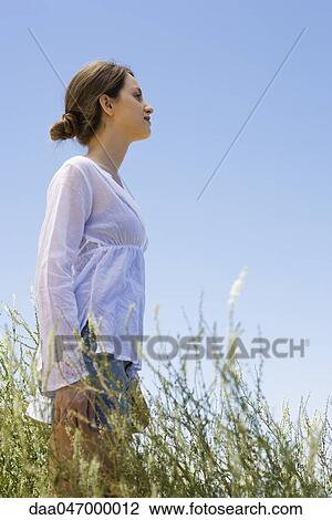 Stock Image - Young woman standing in tall grass, side view. Fotosearch