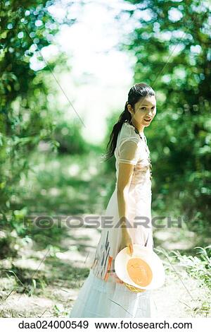 Young woman standing in woods, holding sunhat, smiling at camera View Large Photo Image Stock Photo - Young woman standing in woods, holding sunhat, smiling at camera. Fotosearch