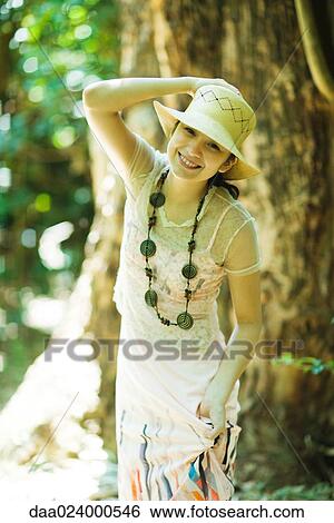 Young woman standing in woods, holding sunhat on head, smiling at camera View Large Photo Image Stock Photograph - Young woman standing in woods, holding sunhat on head, smiling at camera. Fotosearch
