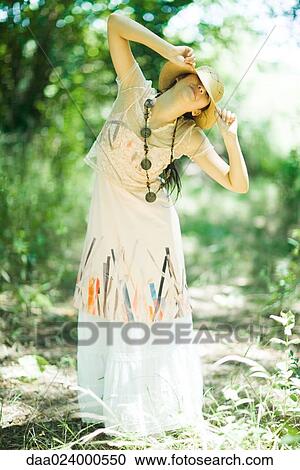 Stock Image - Young woman standing in woods, pulling sunhat over eyes. Fotosearch