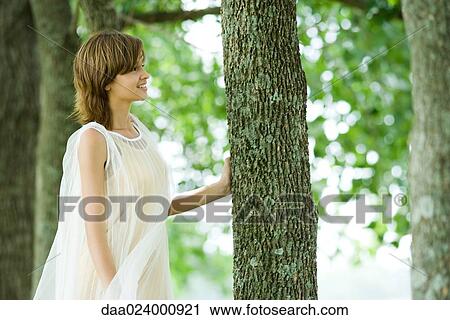 Stock Image - Young woman standing in woods, touching tree trunk, side view. Fotosearch