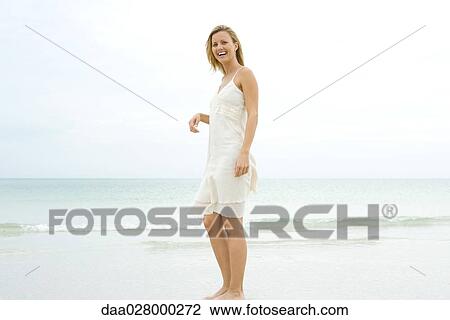 Stock Image - Young woman standing on beach in sundress, laughing. Fotosearch