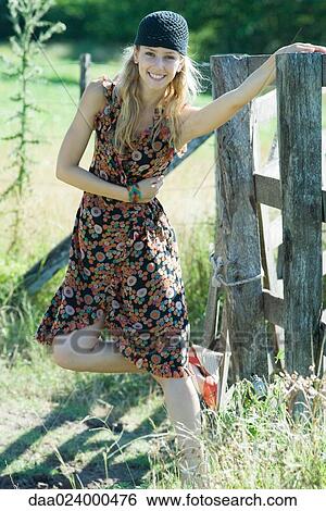 Stock Photograph - Young woman standing on one leg and holding on to rural fence, smiling at camera. Fotosearch