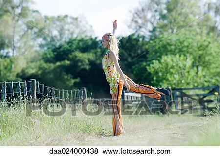 Stock Photo - Young woman standing on one leg in rural field with arm raised. Fotosearch
