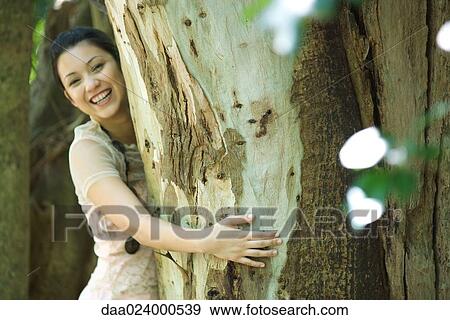 Stock Photo - Young woman standing with arms around tree trunk, smiling at camera. Fotosearch