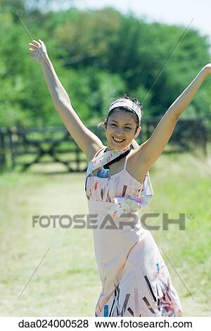 Stock Photo - Young woman standing with arms raised in rural setting, smiling at camera. Fotosearch