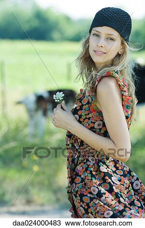 Stock Image - Young woman standing with flower in hand in rural setting, smiling at camera. Fotosearch