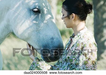 Stock Photo - Young woman touching horse, side view, close-up. Fotosearch