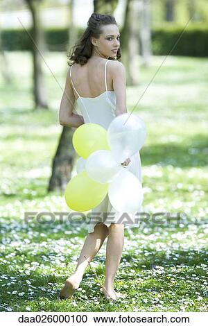 Stock Image - Young woman walking in field of flowers, holding balloons behind back, looking over shoulder at camera. Fotosearch