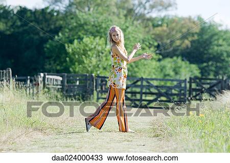 Stock Image - Young woman walking in rural field, smiling at camera, side view. Fotosearch