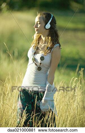 Stock Image - Young woman walking through field, listening to headphones. Fotosearch
