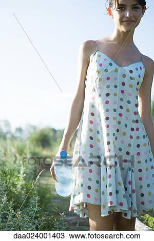 Stock Image - Young woman walking through garden, holding bottle of water, smiling at camera. Fotosearch