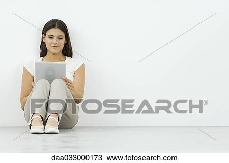 Stock Image - Young woman watching portable DVD player, sitting on the floor, leaning against wall. Fotosearch