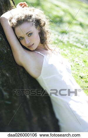 Young woman wearing slip, leaning against tree trunk, arm raised, smiling at camera View Large Photo Image Stock Photograph - Young woman wearing slip, leaning against tree trunk, arm raised, smiling at camera. Fotosearch