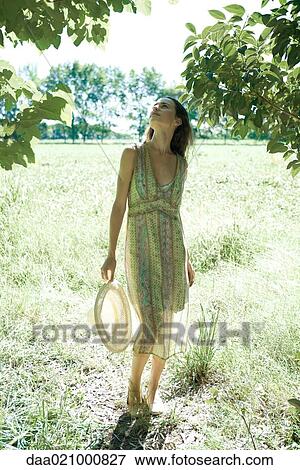 Stock Photo - Young woman wearing sundress and holding sun hat, standing looking up at trees. Fotosearch