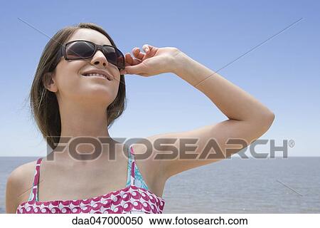 Young woman wearing sunglasses, looking up and smiling, sea in background View Large Photo Image Stock Image - Young woman wearing sunglasses, looking up and smiling, sea in background. Fotosearch
