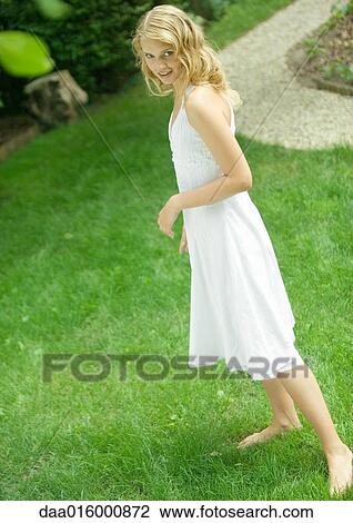 Stock Image - Young woman wearing white dress, standing barefoot on grass. Fotosearch