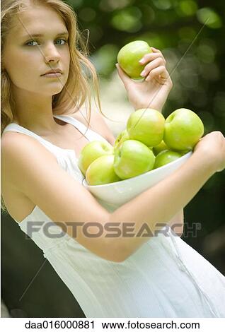 Stock Image - Young woman wearing white dress, holding bowl of apples. Fotosearch