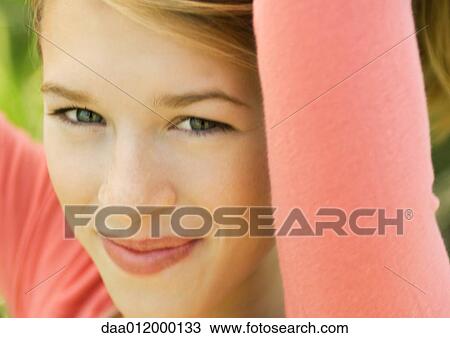 Stock Image - Young woman with arms raised, smiling at camera, close-up. Fotosearch