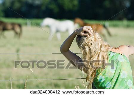 Stock Image - Young woman with hand in hair looking at horses in rural field, rear view. Fotosearch