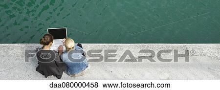 Stock Photo - Young women sitting beside canal, using laptop computer together. Fotosearch