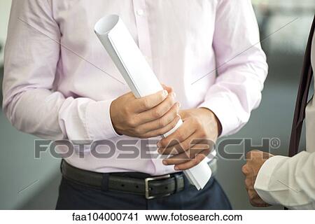 Stock Image - Businessman holding rolled up blueprint, talking with colleague, cropped. Fotosearch