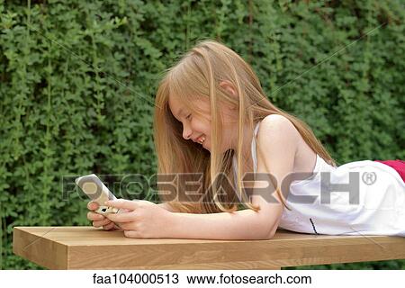 Stock Image - Girl lying on stomach outdoors, playing handheld video game. Fotosearch