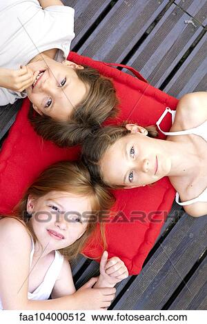 Stock Image - Girls lying down outdoors with heads resting on shared cushion. Fotosearch