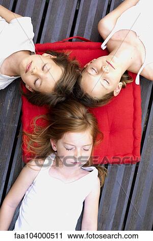 Stock Image - Girls napping outdoors with heads resting on shared cushion. Fotosearch