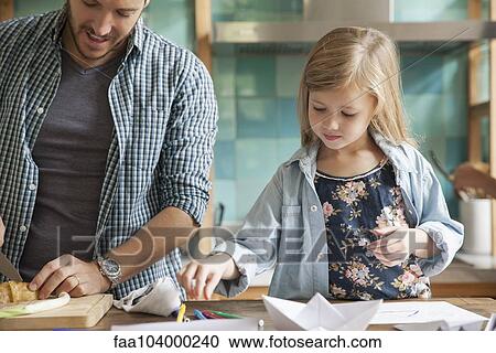 Stock Image - Little girl drawing in kitchen while family prepares meal. Fotosearch