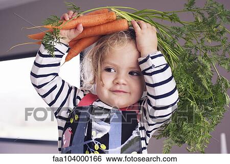 Stock Photograph - Little girl holding fresh carrots over her head, portrait. Fotosearch