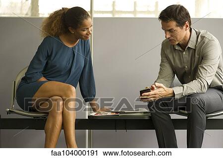 Stock Image - Professionals chatting in waiting room, man showing woman smartphone. Fotosearch