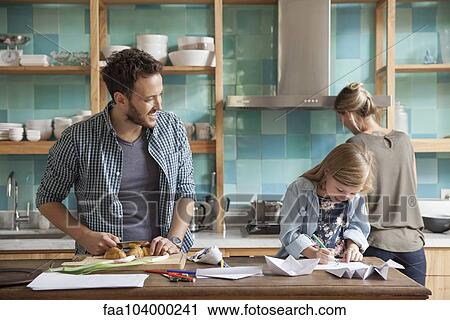 Stock Image - Young daughter drawing ar kitchen counter while parents prepare meal. Fotosearch