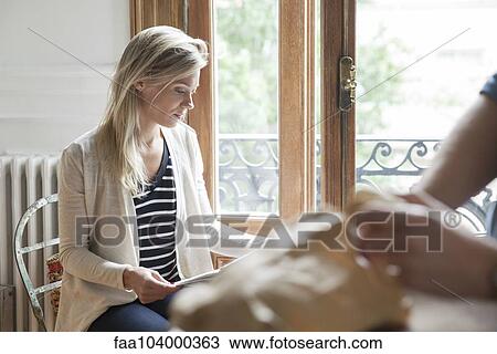 Stock Image - Young woman sitting by window reading. Fotosearch