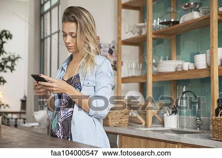 Young woman standing in kitchen with cell phone View Large Photo Image Stock Photo - Young woman standing in kitchen with cell phone. Fotosearch