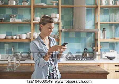 Stock Photo - Young woman standing in kitchen with cell phone. Fotosearch