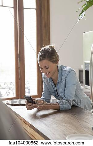 Stock Image - Young woman using smartphone in kitchen. Fotosearch