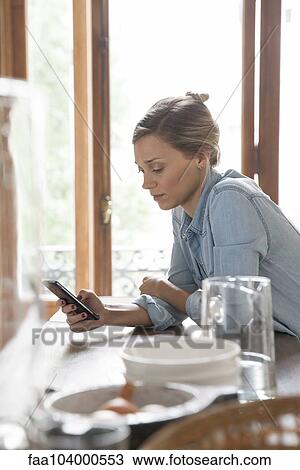 Stock Image - Young woman using smartphone in kitchen. Fotosearch