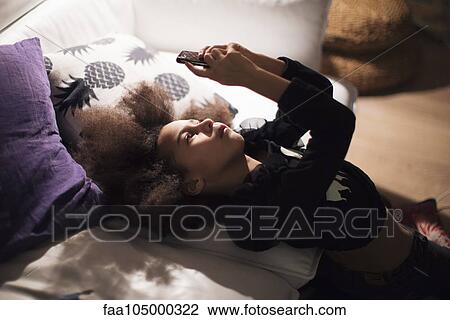 Stock Image - Girl reclining against sofa, engrossed in smartphone. Fotosearch