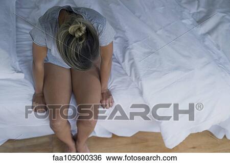 Stock Photograph - Woman sitting on edge of bed with head lowered. Fotosearch