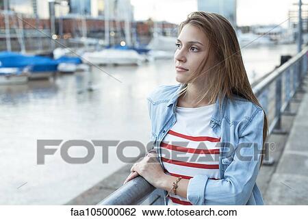 Stock Image - Young woman leaning against railing, looking at view. Fotosearch