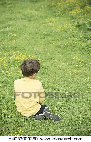 Stock Image - Boy sitting alone in grass, rear view. Fotosearch