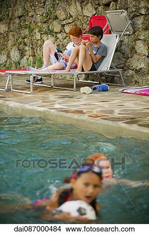 Boys playing handheld video games beside swimming pool View Large Photo Image Picture - Boys playing handheld video games beside swimming pool. Fotosearch