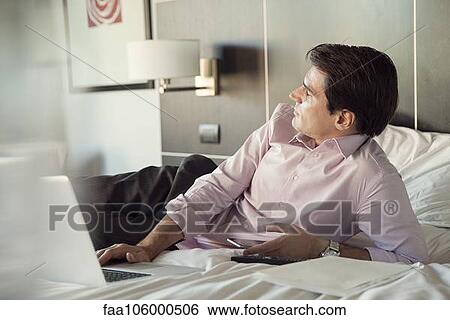 Businessman lying on hotel bed with cell phone and laptop computer, looking away in thought View Large Photo Image Stock Photograph - Businessman lying on hotel bed with cell phone and laptop computer, looking away in thought. Fotosearch