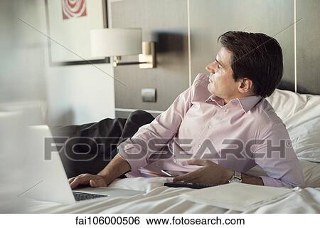 Businessman lying on hotel bed with cell phone and laptop computer, looking away in thought View Large Photo Image Stock Photograph - Businessman lying on hotel bed with cell phone and laptop computer, looking away in thought. Fotosearch