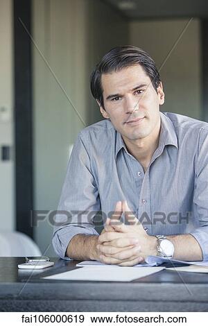Stock Photo - Businessman sitting at desk with clasped hands, portrait. Fotosearch