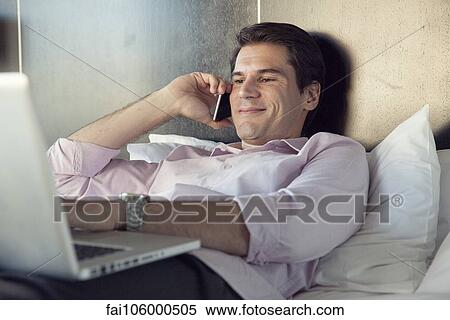 Stock Photography - Businessman using cell phone and laptop computer in bed. Fotosearch