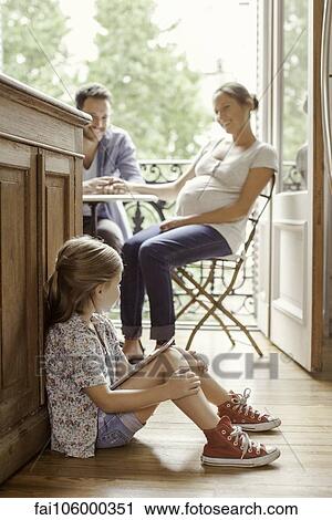 Stock Image - Girl sitting on floor with digital tablet as parents chat in background. Fotosearch